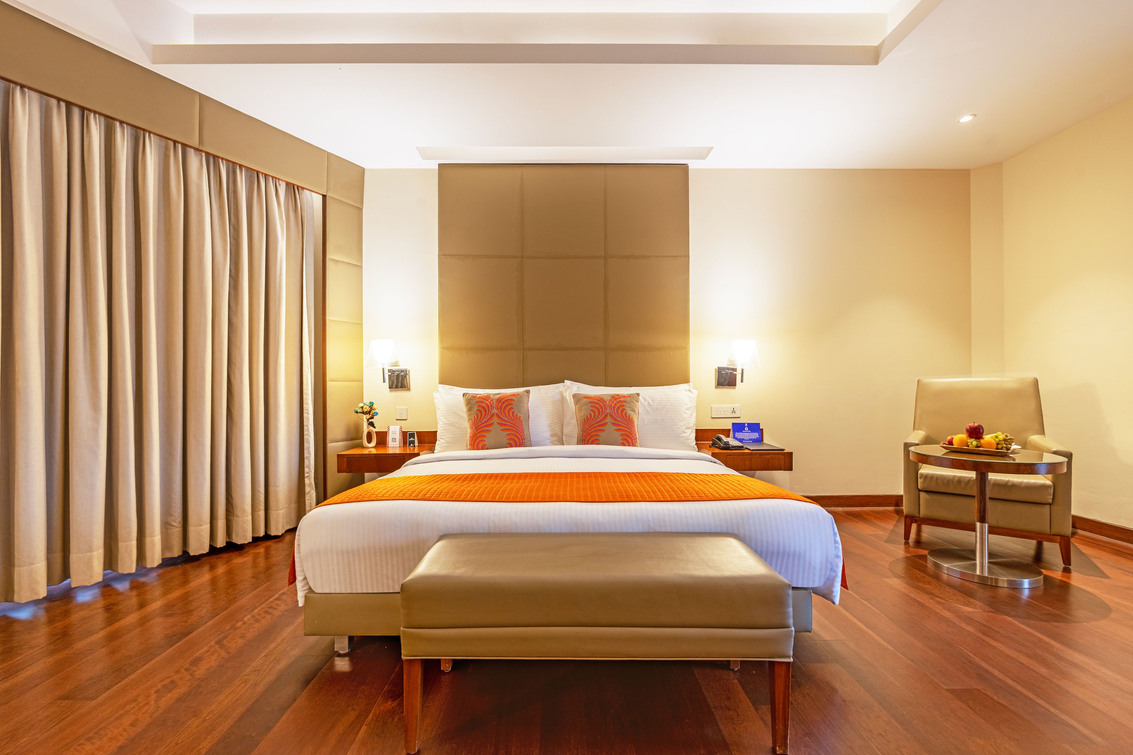 A hotel room with a large bed, a bedside table with a lamp, a chair, and a small table with fruit.