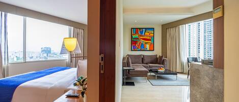 A hotel room with a bed, a desk, a coffee table, and a sofa. There is a painting on the wall and a view of the city through the window.