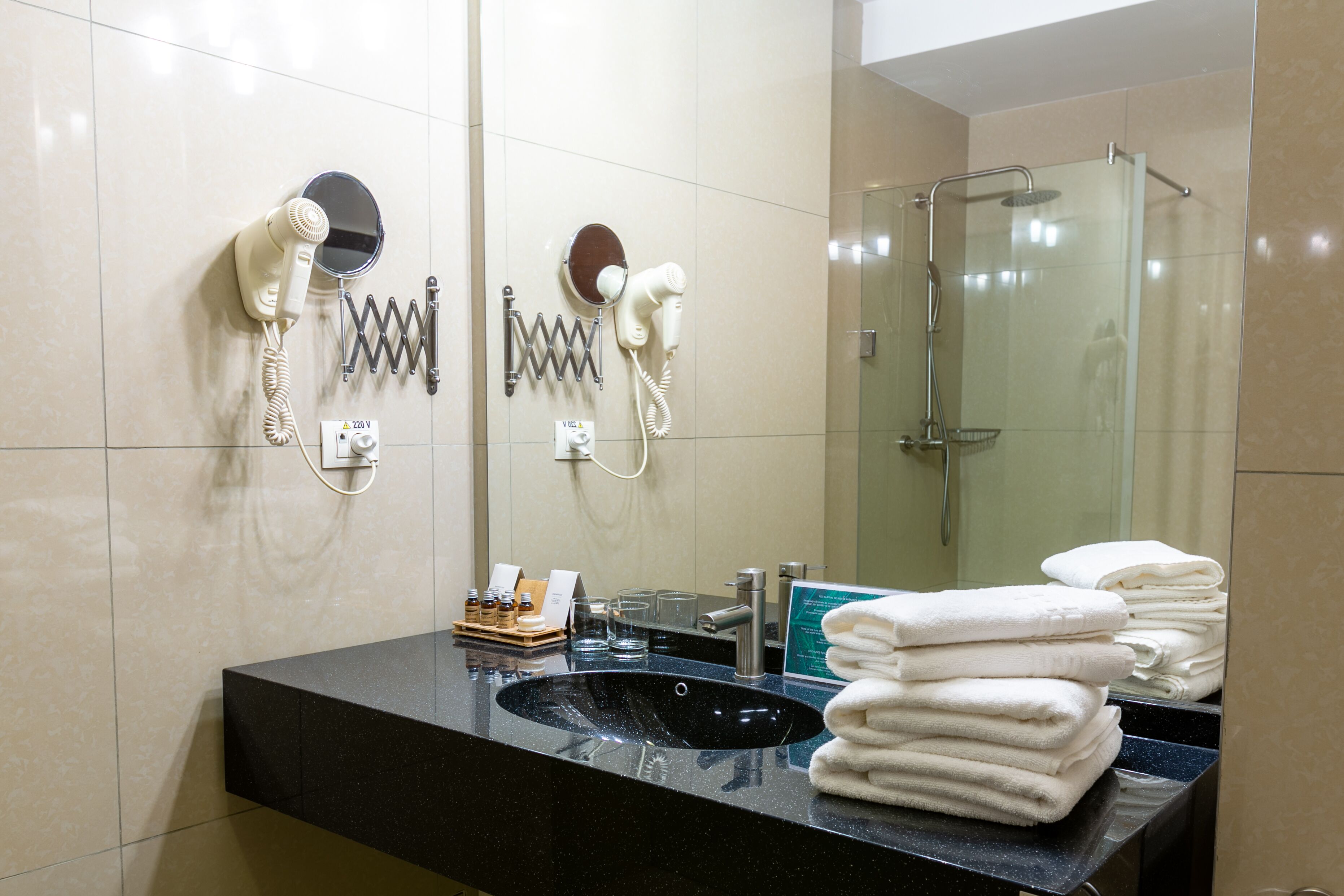 A bathroom with a black sink, two hair dryers mounted on the wall, a glass shower enclosure, and a stack of towels on the counter.