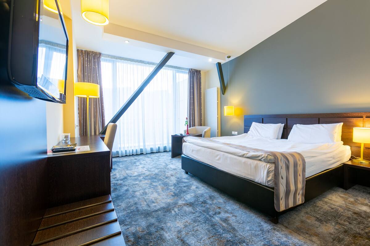 A hotel room with a large bed, a desk, a chair, a TV, and a window with curtains.
