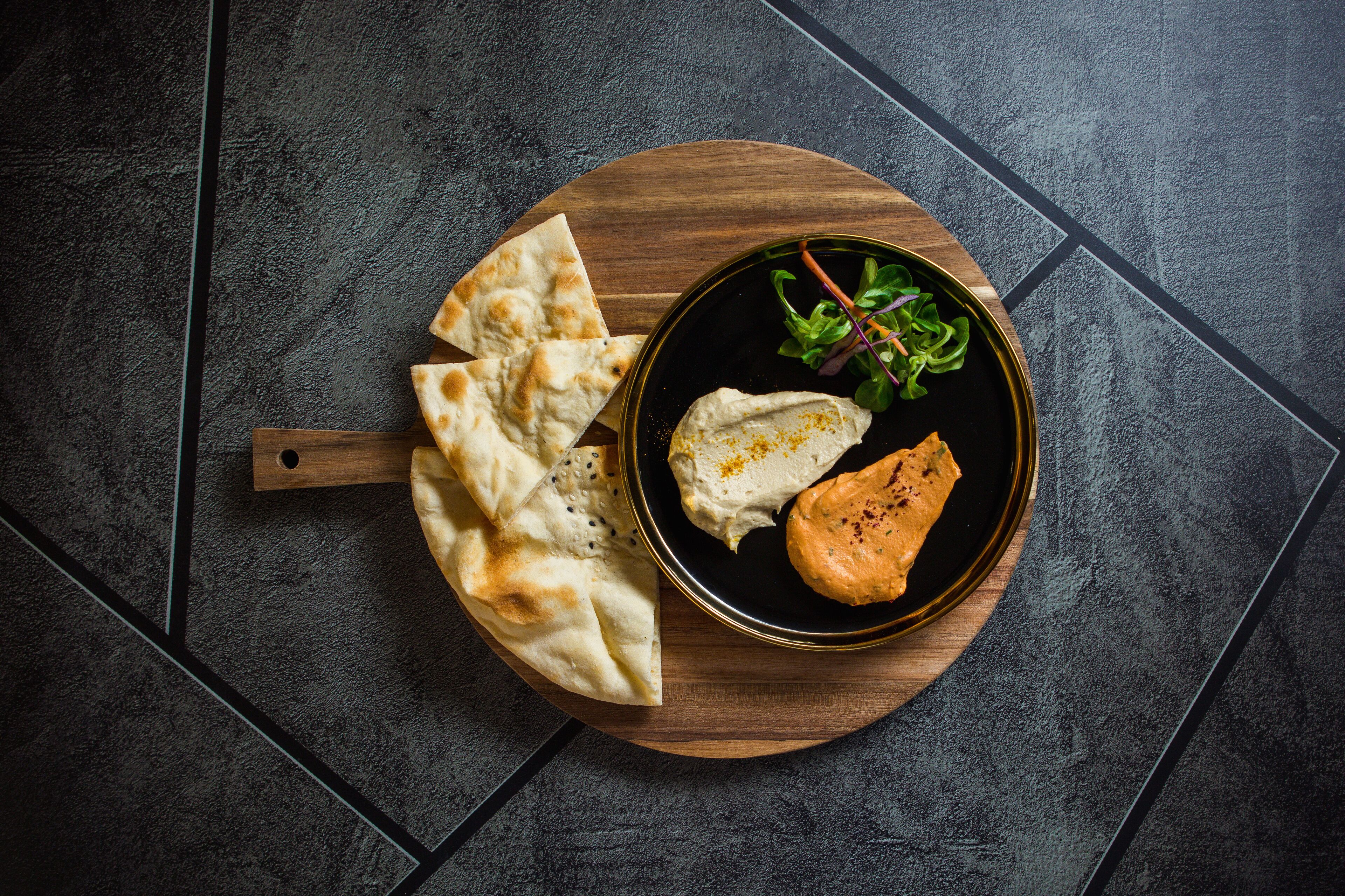 Une planche en bois garnie de galettes, un bol de houmous et un morceau de poulet grillé, le tout posé sur un sol carrelé foncé.
