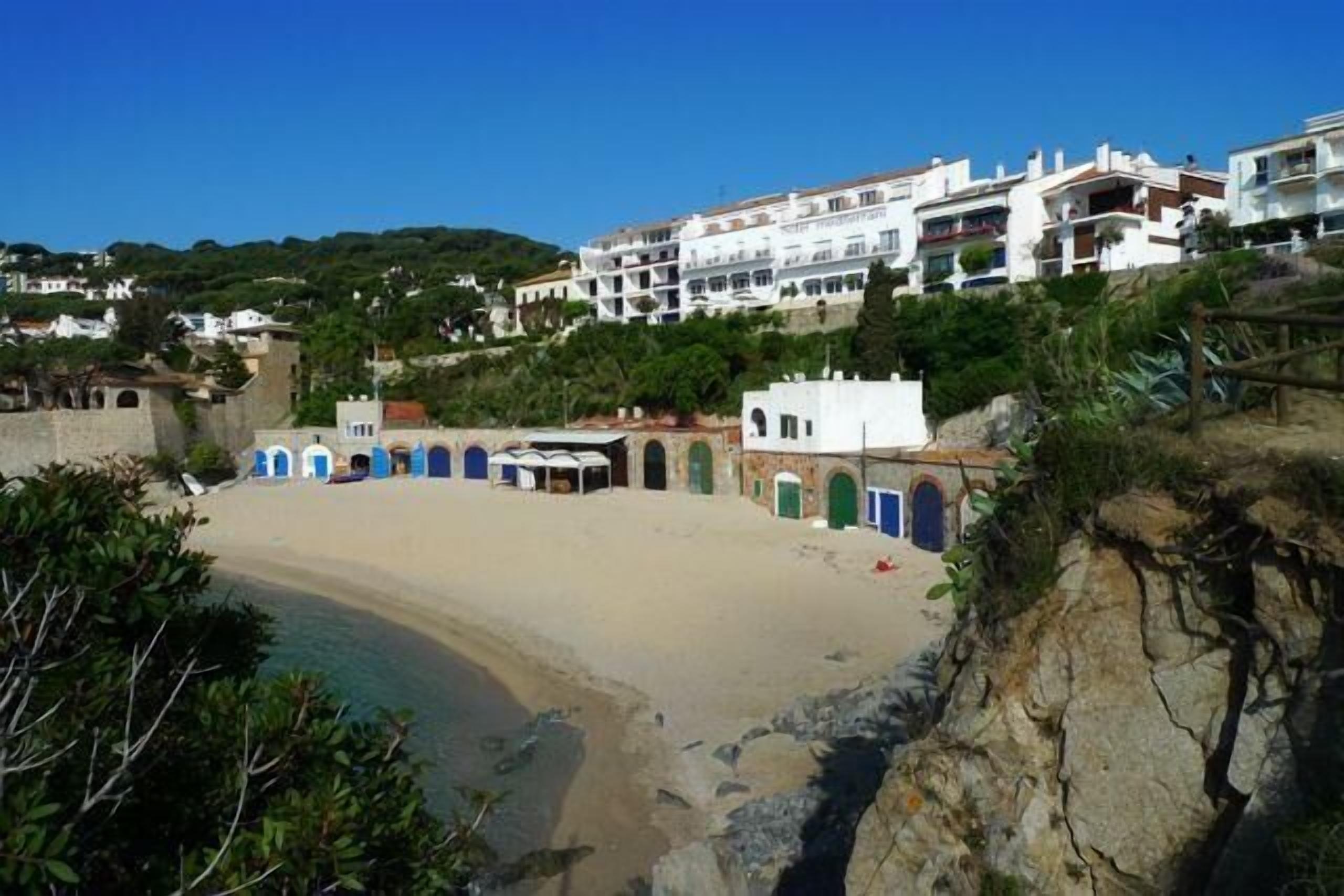 A beach with a curved shoreline, a small building with blue doors, and white buildings in the background.