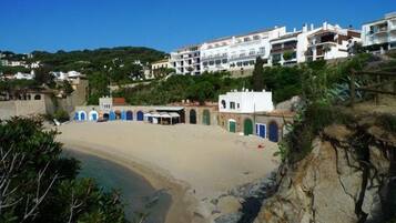 Una playa con una costa curva, un pequeño edificio con puertas azules y edificios blancos al fondo.