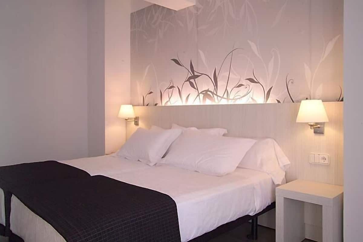 Un lit bien fait, avec du linge de lit blanc et une couverture sombre, flanqué de deux tables de chevet sur lesquelles sont posées des lampes.