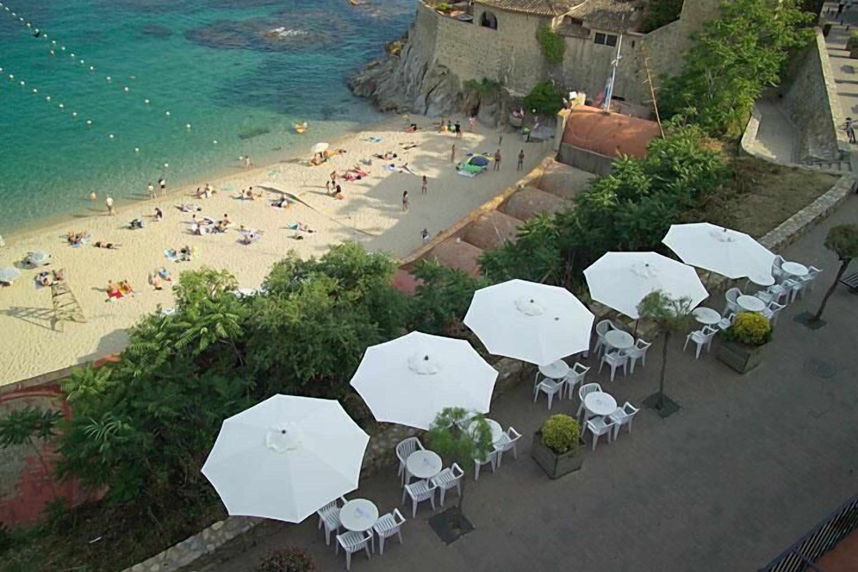 An aerial view of a beach with umbrellas and tables, a rocky coastline, and a building with a thatched roof.