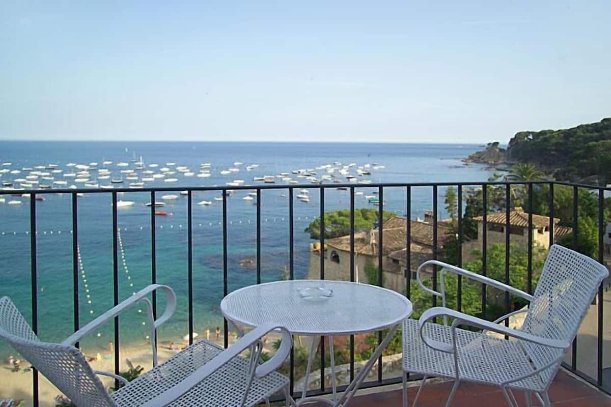 Un balcon avec deux chaises en métal blanc et une petite table ronde, donnant sur une zone côtière où se trouvent de nombreux bateaux et une côte rocheuse.