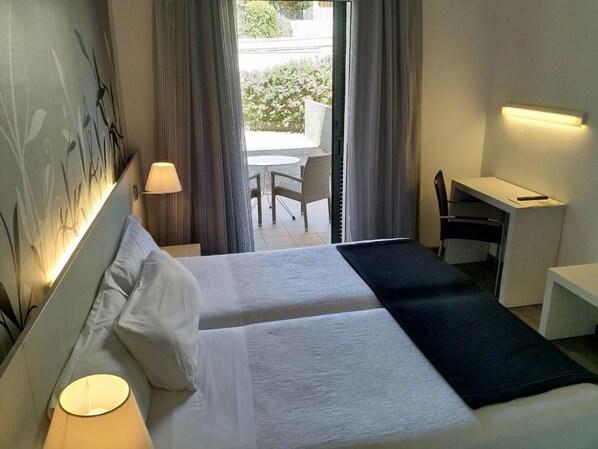 A hotel room with two beds, a desk, a chair, and a view of the outdoors. - Hotel Mediterrani (Palafrugell)