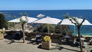 Un espacio al aire libre con mesas y sillas, sombrillas blancas y vistas al mar y un yate.