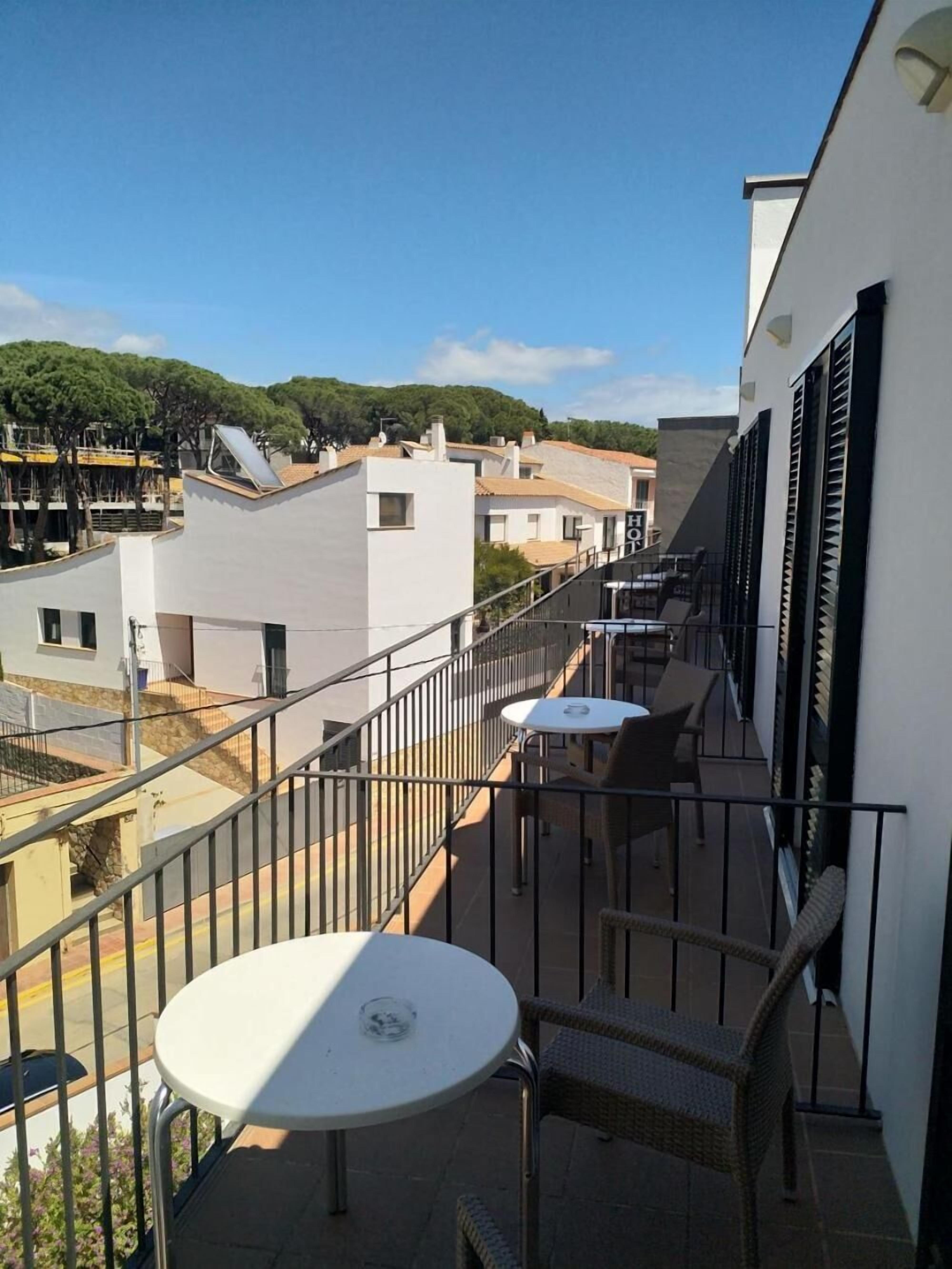 A balcony with a white table and wicker chairs, overlooking a residential area with white buildings and a clear sky.