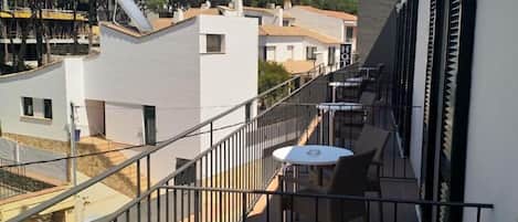 A balcony with a white table and wicker chairs, overlooking a residential area with white buildings and a clear sky.