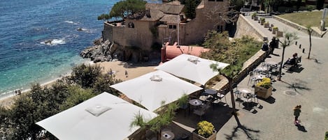 Una zona costera con un edificio de piedra, mesas al aire libre con sombrillas y vistas al mar.