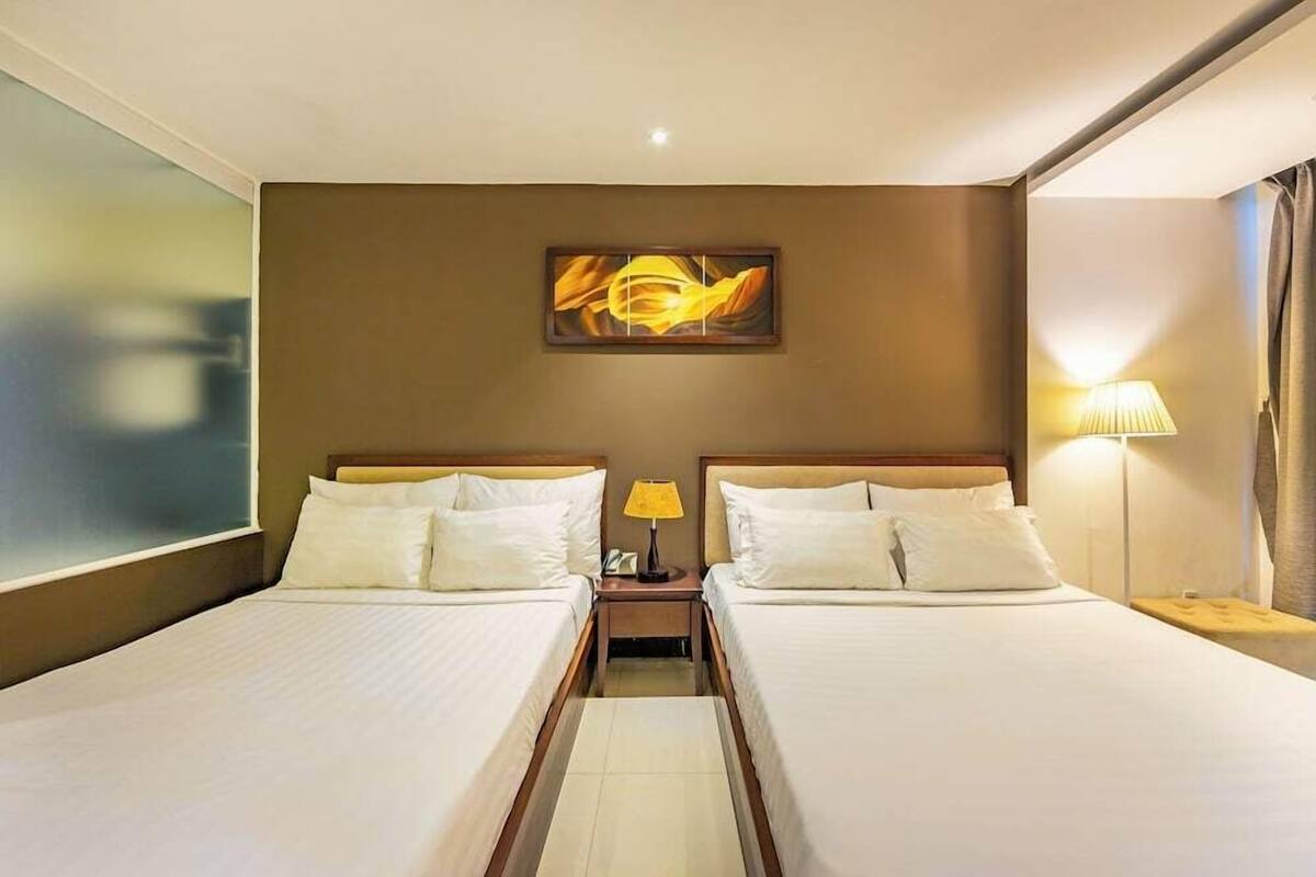 A hotel room with two beds, a painting on the wall, a nightstand with a lamp, and a window with curtains.