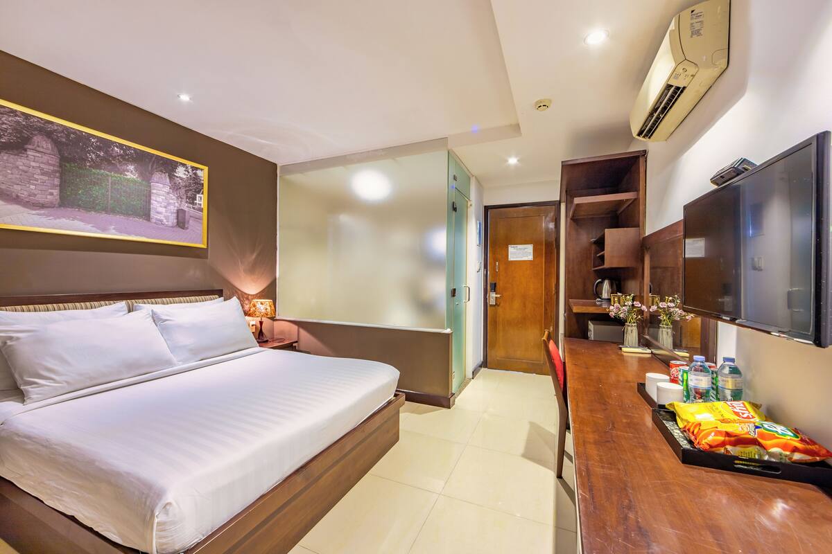 A hotel room with a bed, a desk with a tray of snacks, a TV, and a painting on the wall.