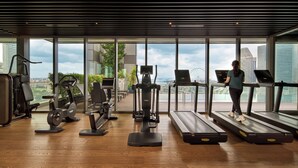 Une salle de sport équipée de tapis de course, de vélos d’exercice et de poids, avec de grandes fenêtres offrant une vue sur la ville.