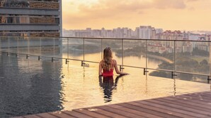 Une femme est assise sur un balcon en verre, d’où elle a une vue sur la ville au coucher du soleil.