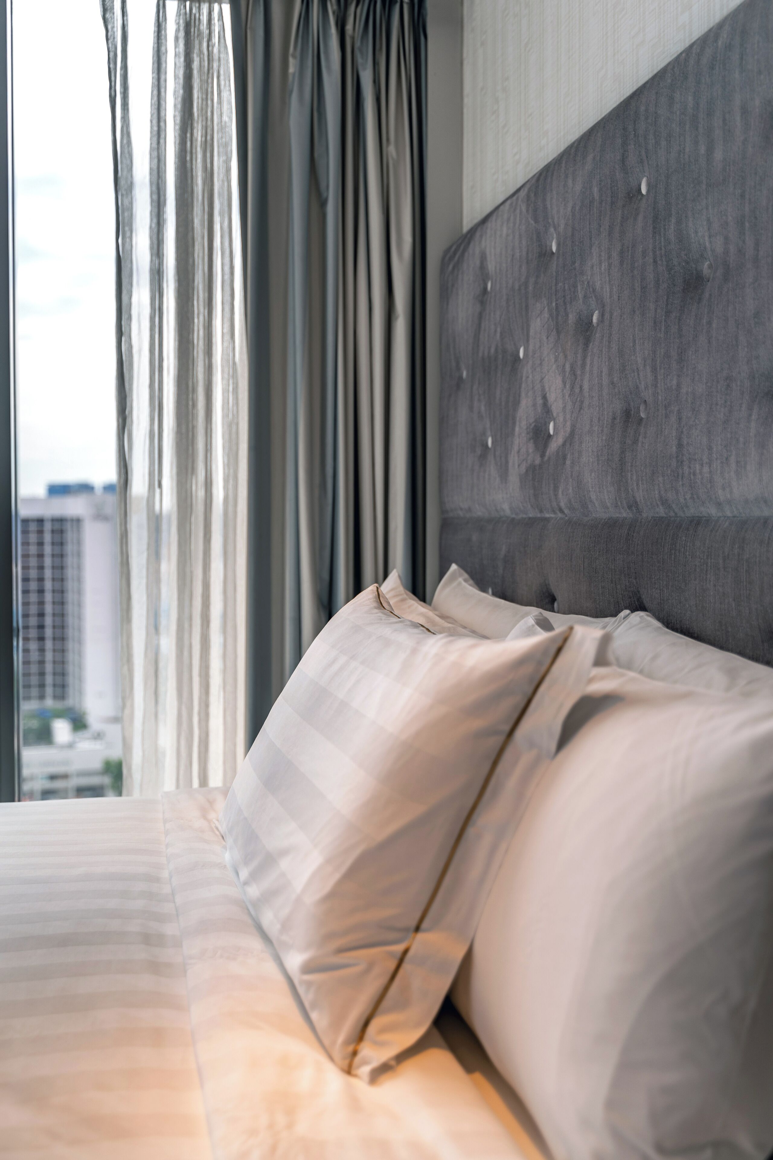 Una habitación de hotel moderna con una cama grande, cabecera gris y vistas a edificios a través de cortinas traslúcidas.
