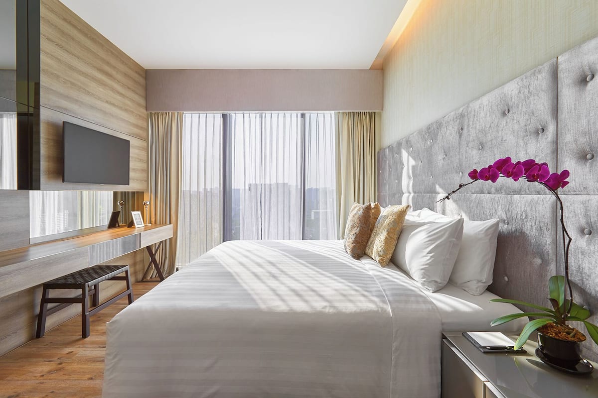 Een moderne hotelkamer met een groot bed, een flatscreen televisie aan de muur, een bureau met stoel en een groot raam met dunne gordijnen.
