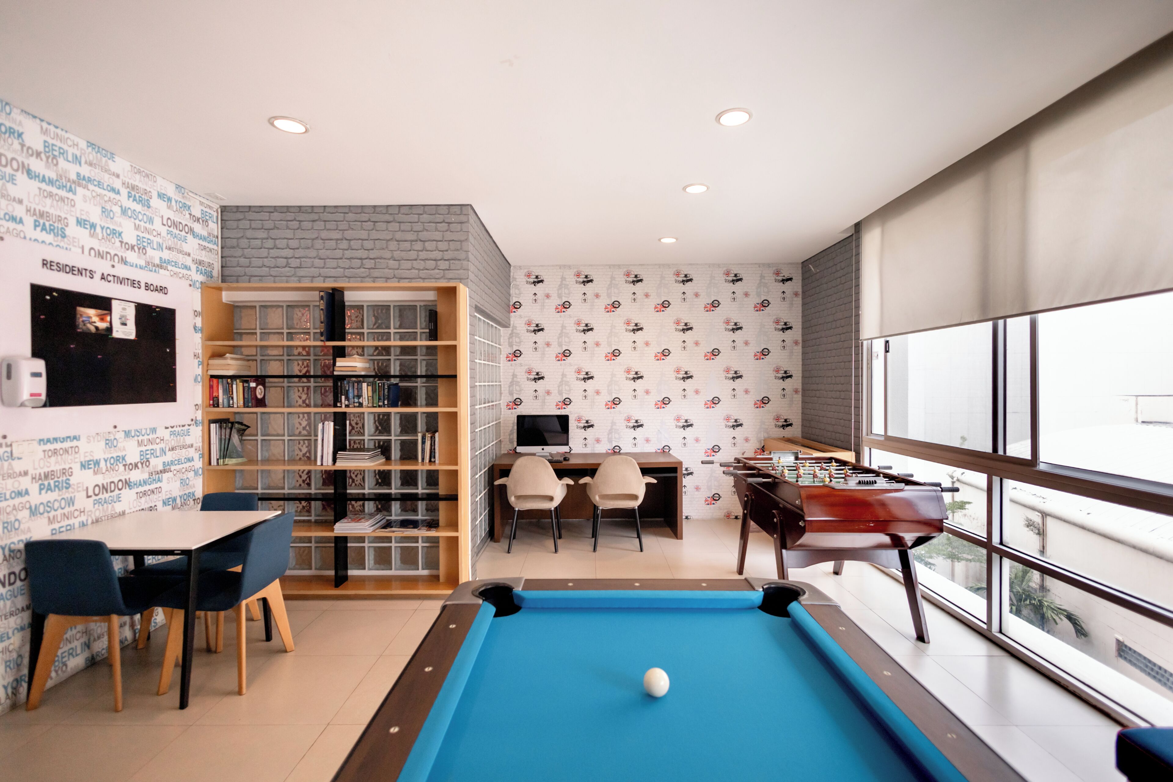 A pool table with a ball, a foosball table, a bookshelf, a desk with two chairs, and a city-themed wall.