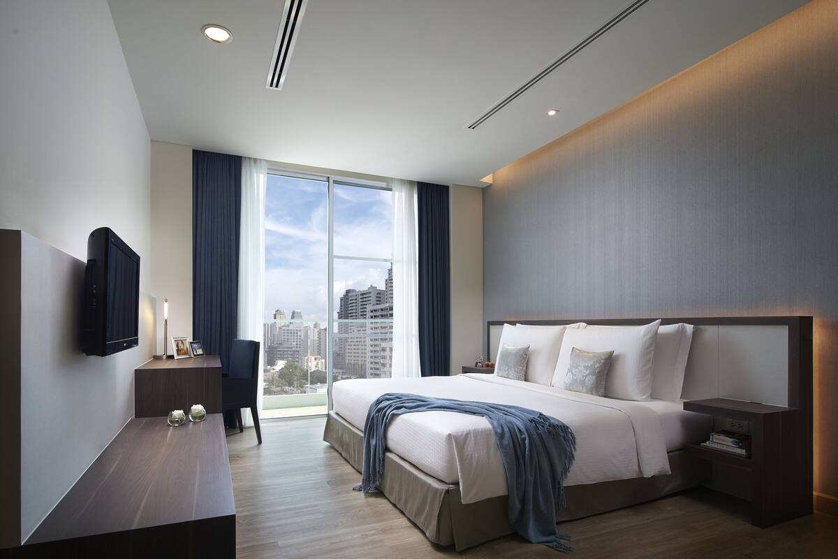 A modern hotel room with a large bed, a flat-screen TV mounted on the wall, a desk with a chair, and a large window offering a city view.