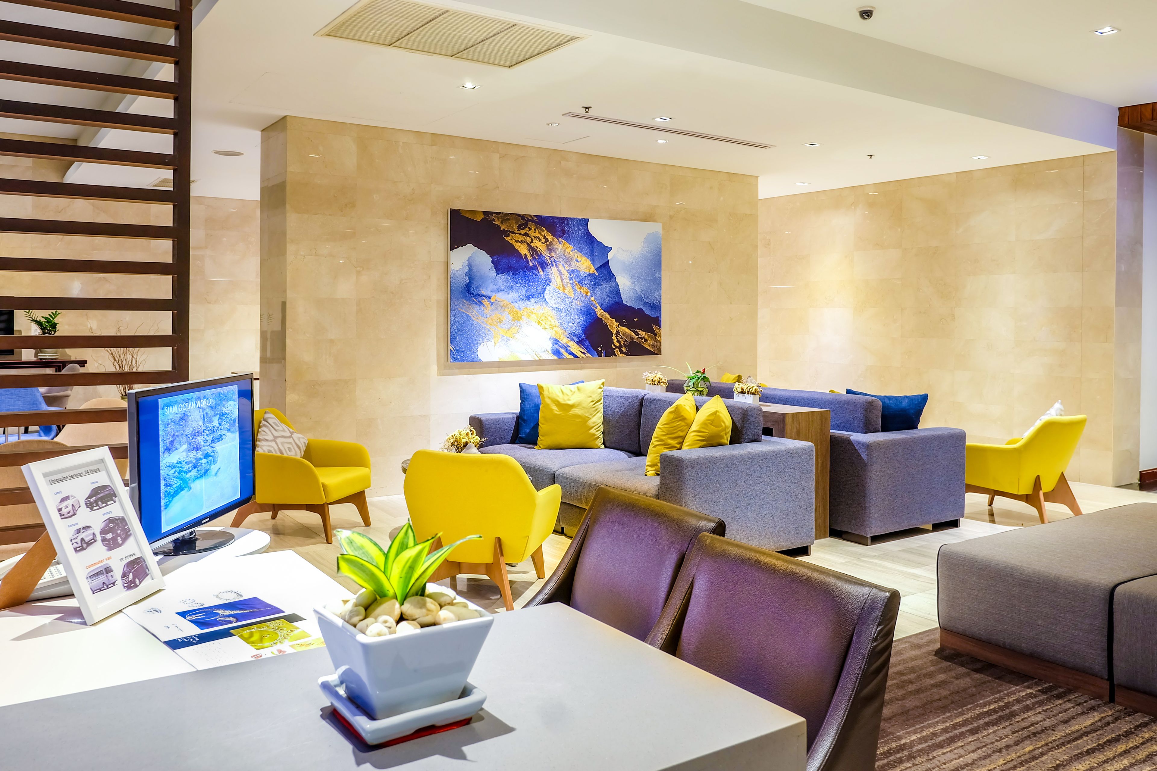 Un hall d’hôtel moderne doté d’un espace détente avec des canapés et des fauteuils, d’un grand tableau abstrait au mur et d’un bureau avec un écran d’ordinateur et des brochures d’information.