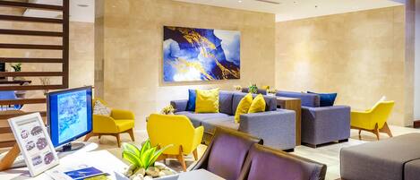 Un hall d’hôtel moderne doté d’un espace détente avec des canapés et des fauteuils, d’un grand tableau abstrait au mur et d’un bureau avec un écran d’ordinateur et des brochures d’information.