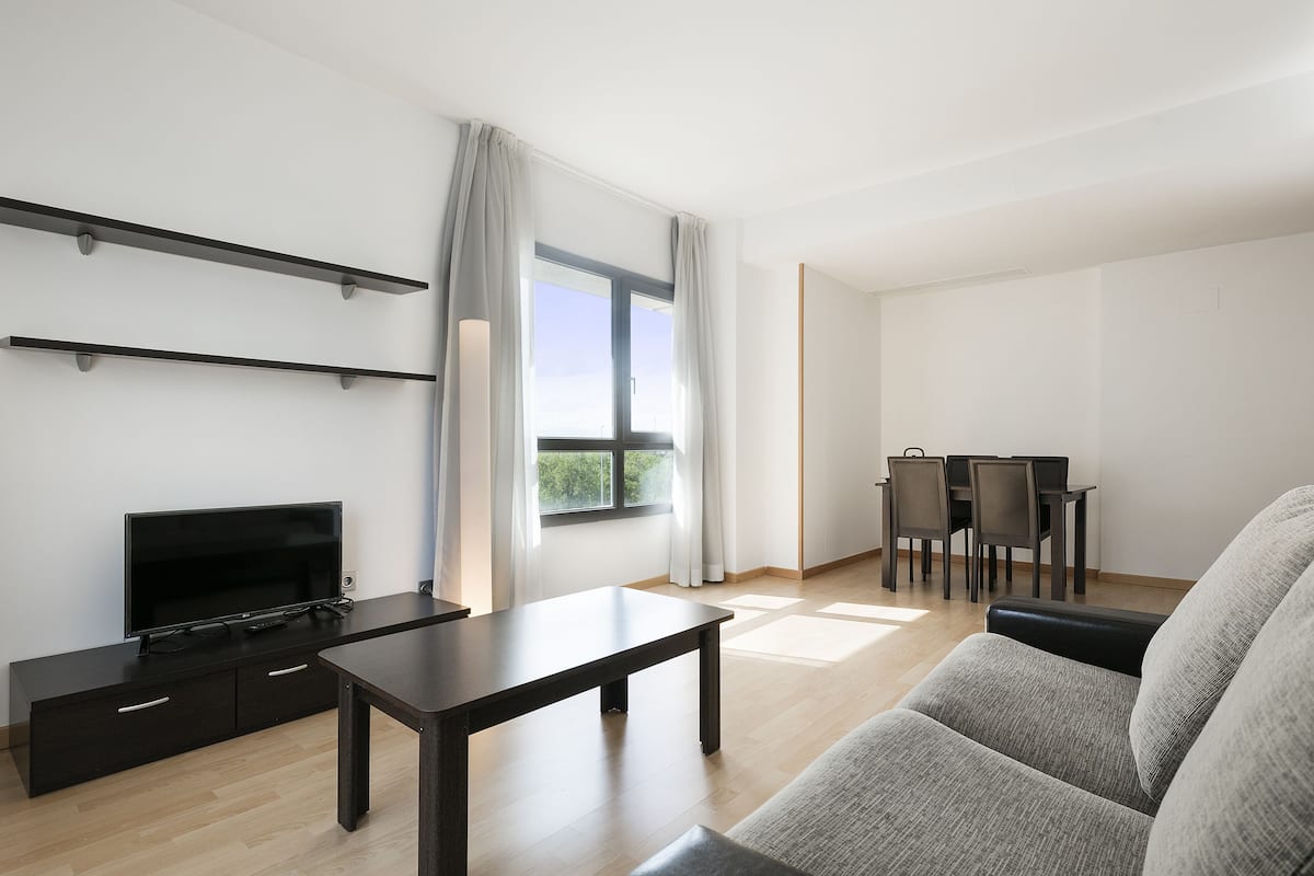 Ein modernes Wohnzimmer mit Fernseher, einem dunkelholzbigen Couchtisch, einem grauen Sofa und einem Essbereich mit Tisch und Stühlen.
