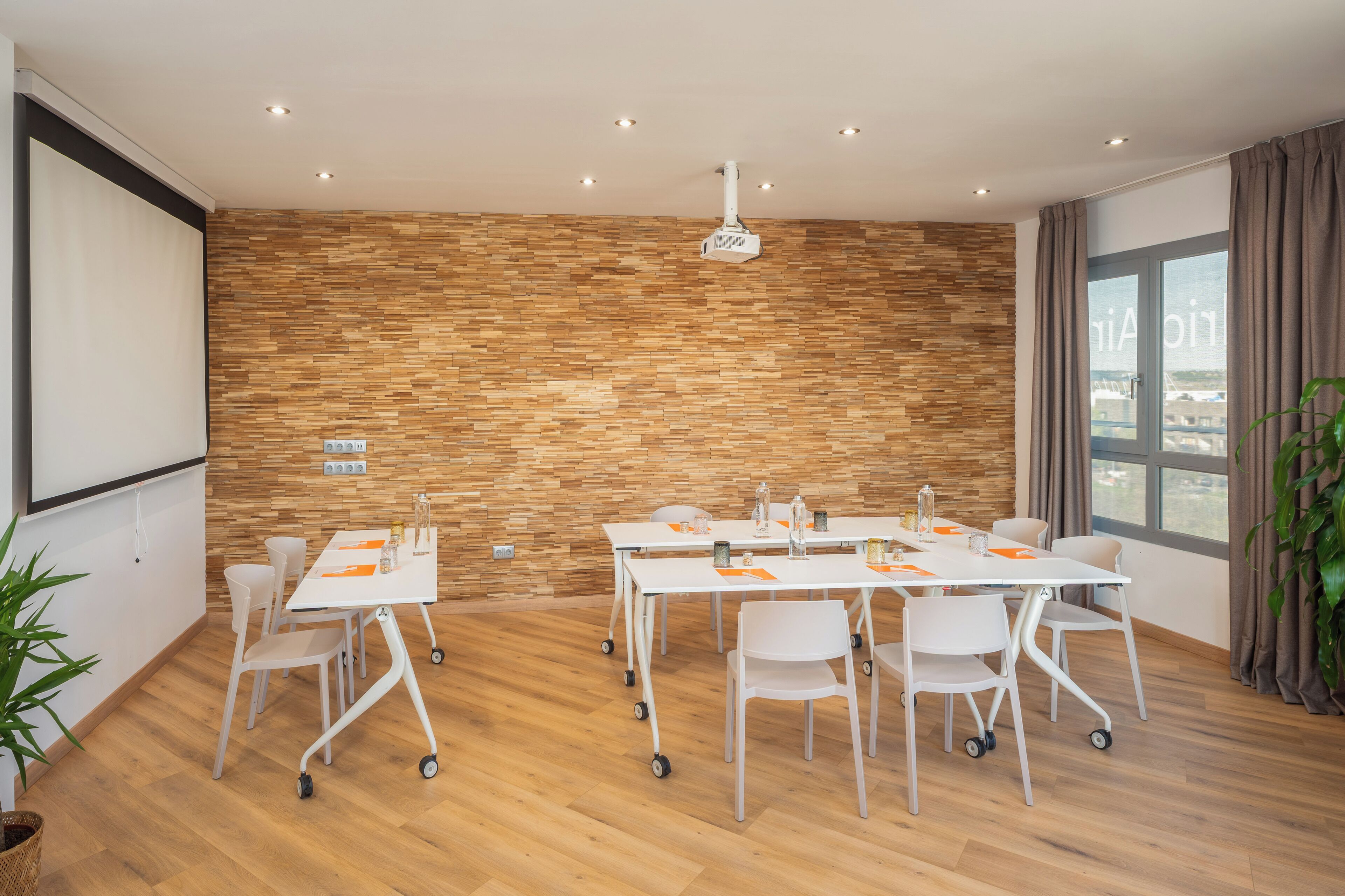 Un salón de conferencias con pared de madera, mesas y sillas blancas, un proyector y una ventana con cortinas.