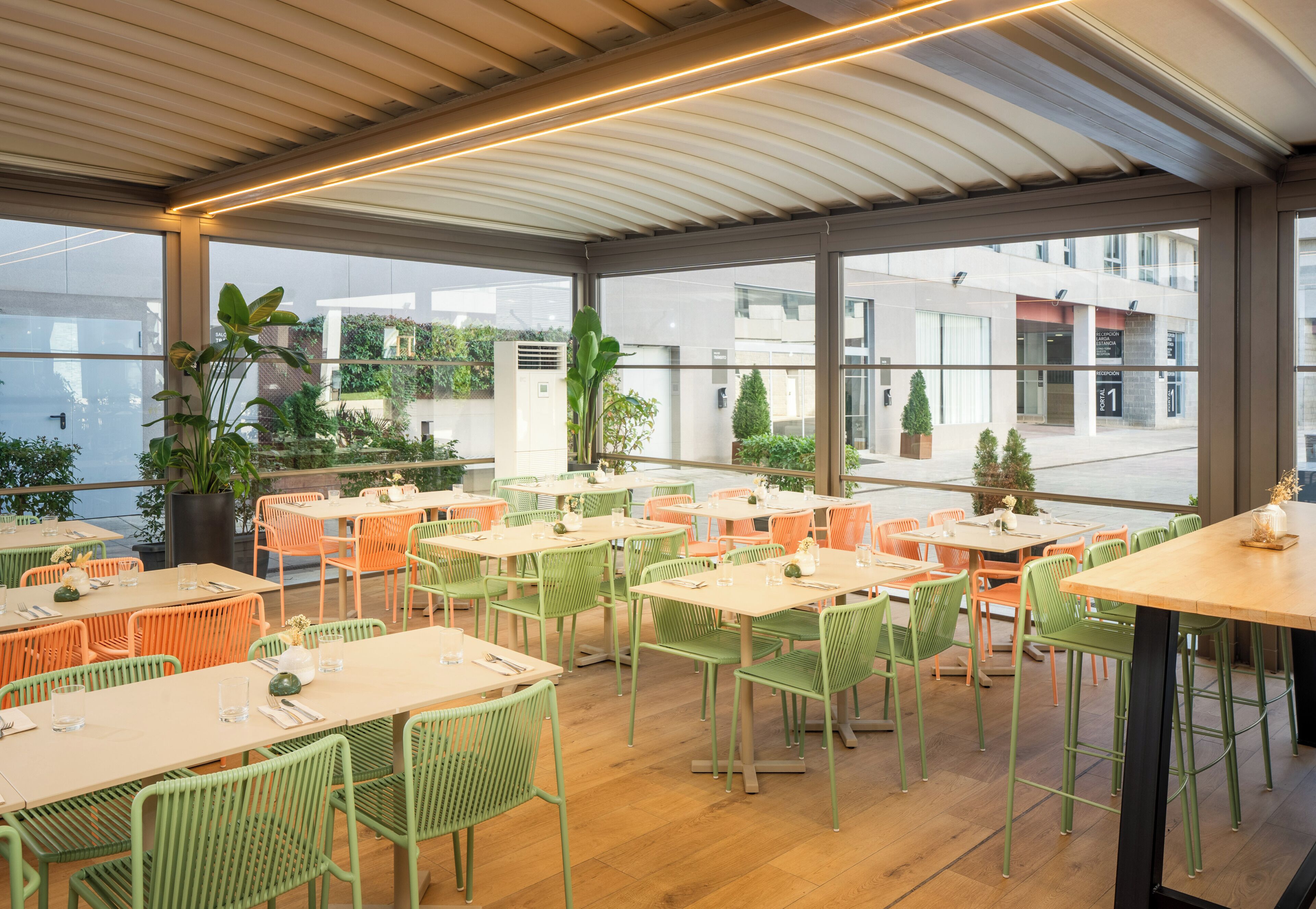 Un comedor interior con mesas y sillas, ventanas amplias y plantas en macetas.