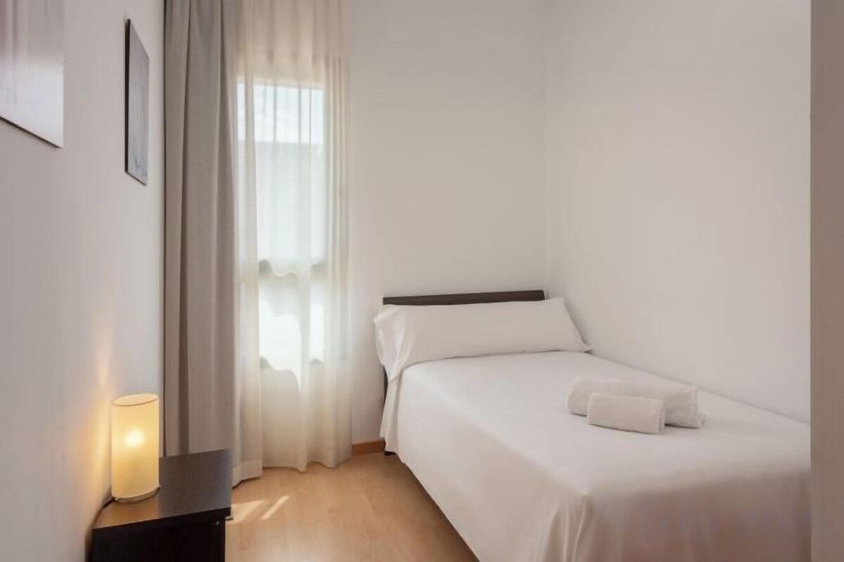Una cama individual con cabecera blanca en habitación con suelo de madera y mesita de noche con lámpara.
