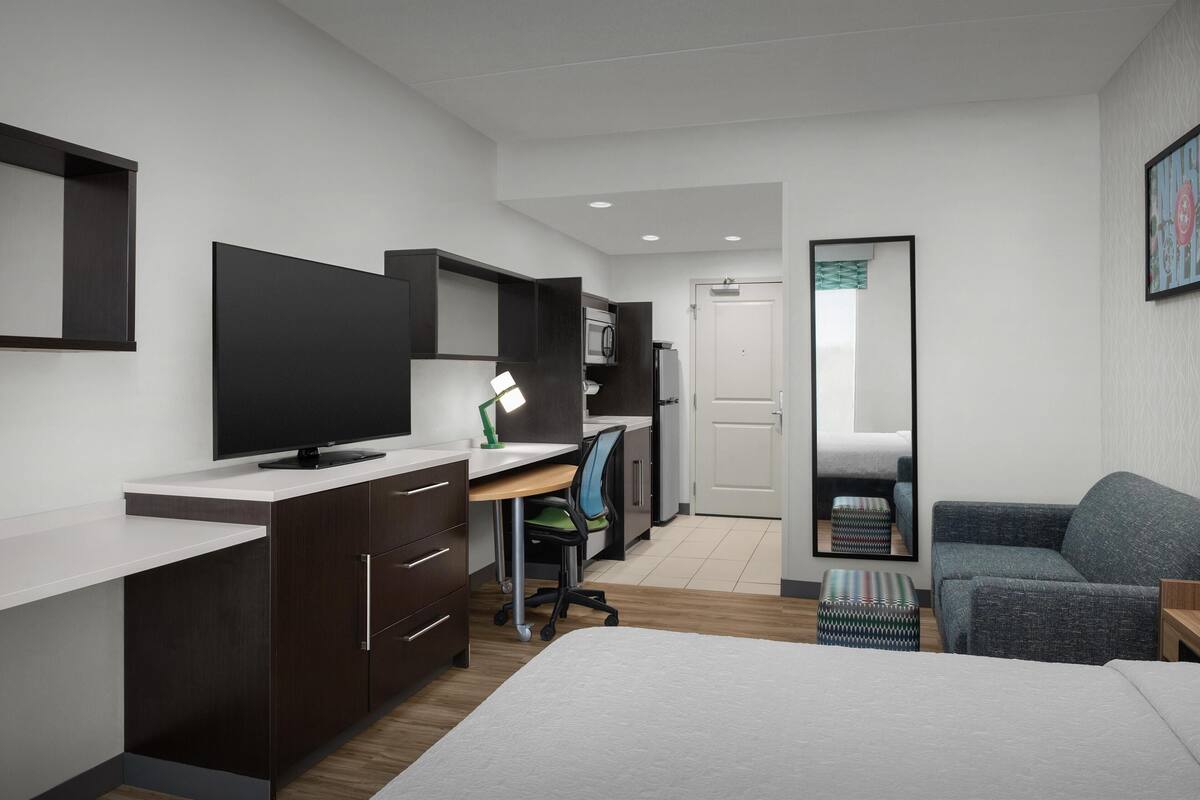 Ein modernes Schlafzimmer mit Bett, Schreibtisch, Stuhl, Fernseher, Mikrowelle und einer Tür zu einem weiteren Raum.