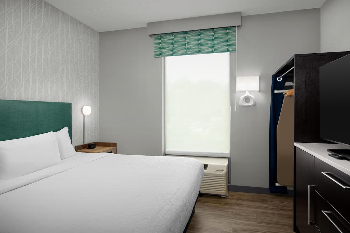 Ein Hotelzimmer mit Bett, Nachttisch, Lampe, Fenster mit Jalousien und einem eingebauten TV-Schrank.