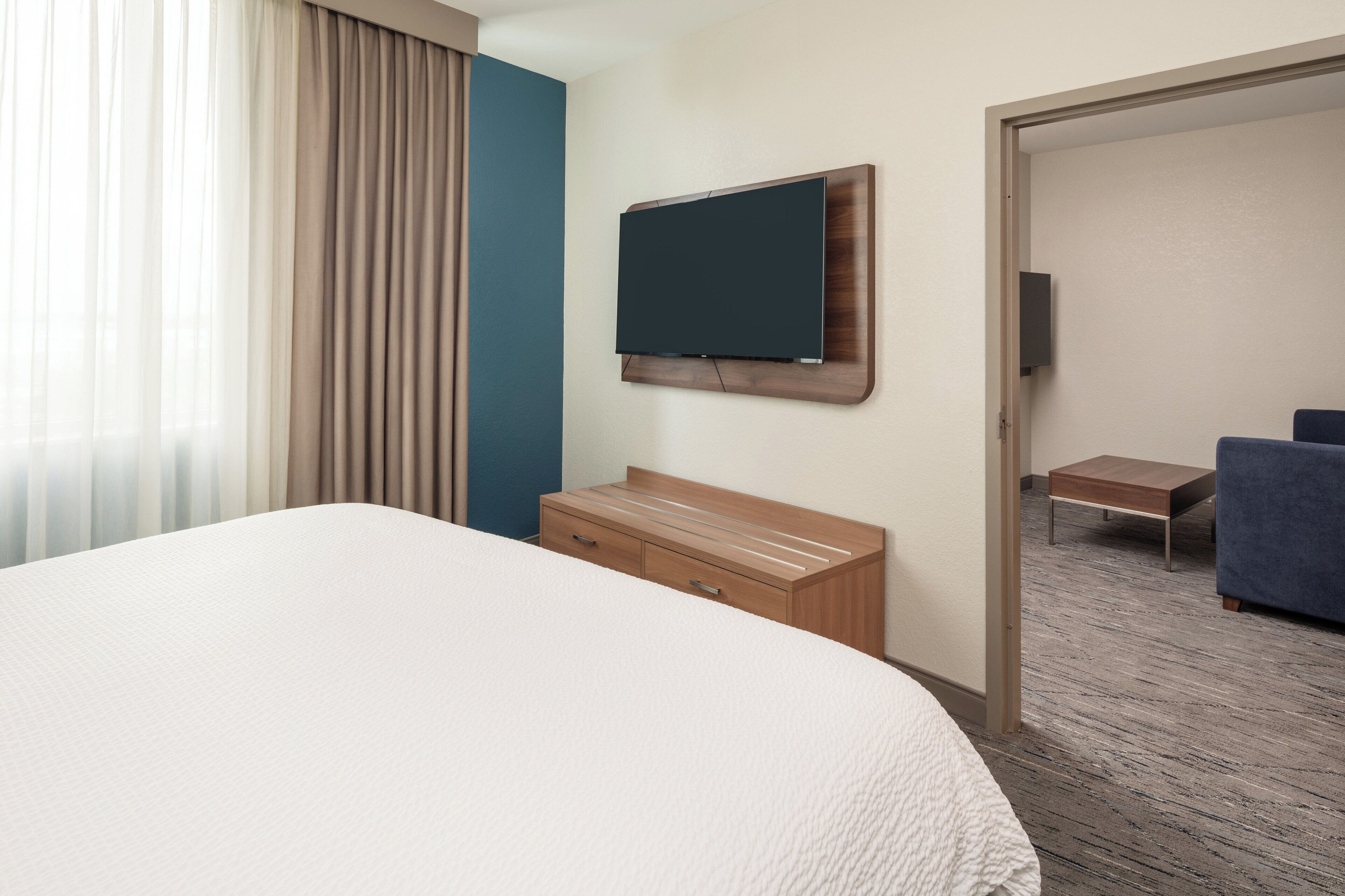 A hotel room with a large bed, a wooden dresser, a flat-screen TV mounted on the wall, a small table, and a blue chair.