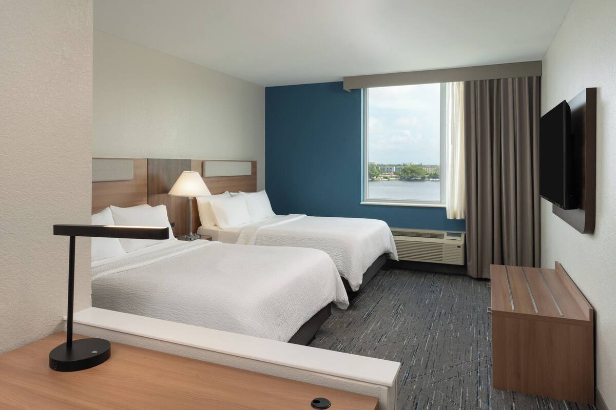 A hotel room with two beds, a TV mounted on the wall, a desk, and a window with curtains.