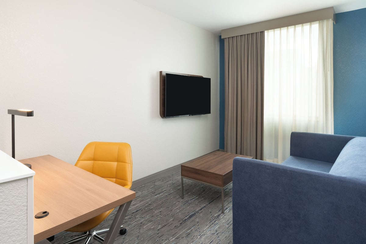 A hotel room with a wooden desk, a yellow chair, a blue sofa, a flat-screen TV mounted on the wall, and a window with curtains.