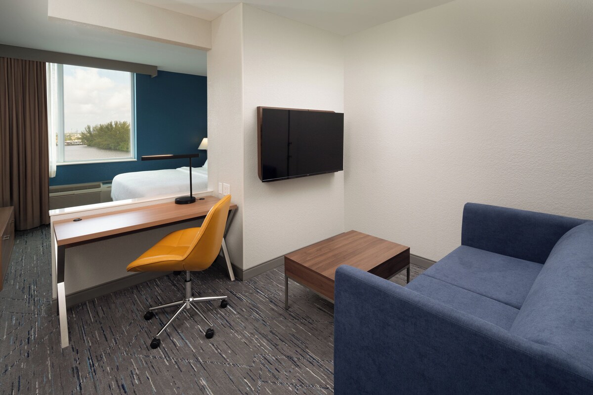 Una habitación de hotel con cama, escritorio y silla, televisor montado en la pared y un sofá azul.