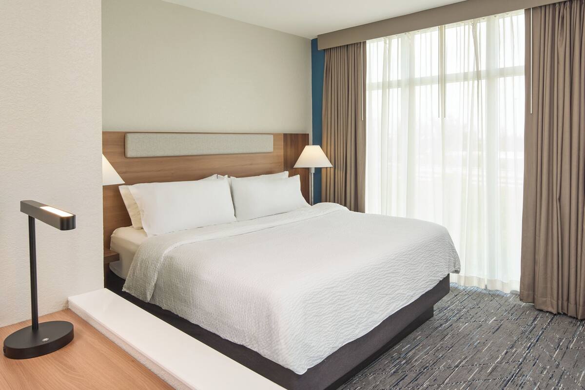 A hotel room with a large bed, a bedside table, a lamp, and a window with curtains.