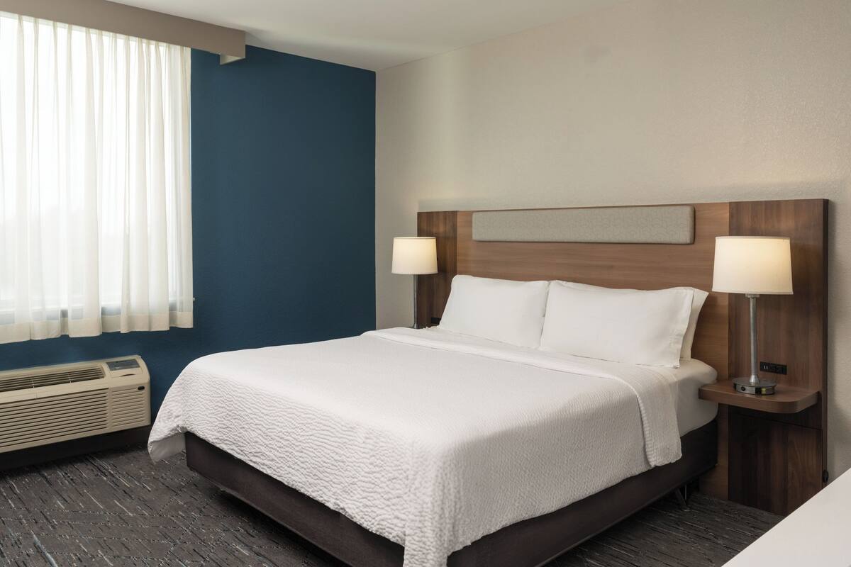 A hotel room with a large bed, two bedside lamps, a headboard with a textured panel, a window with sheer curtains, and a wall-mounted air conditioning unit.