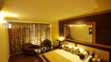 A hotel room with a large bed, a chair, a desk with a lamp, a mirror, and curtains.
