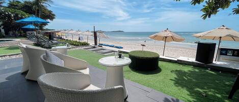 A terrace with wicker furniture, a small table, and a decorative item. There are umbrellas and a view of the beach and ocean in the background.