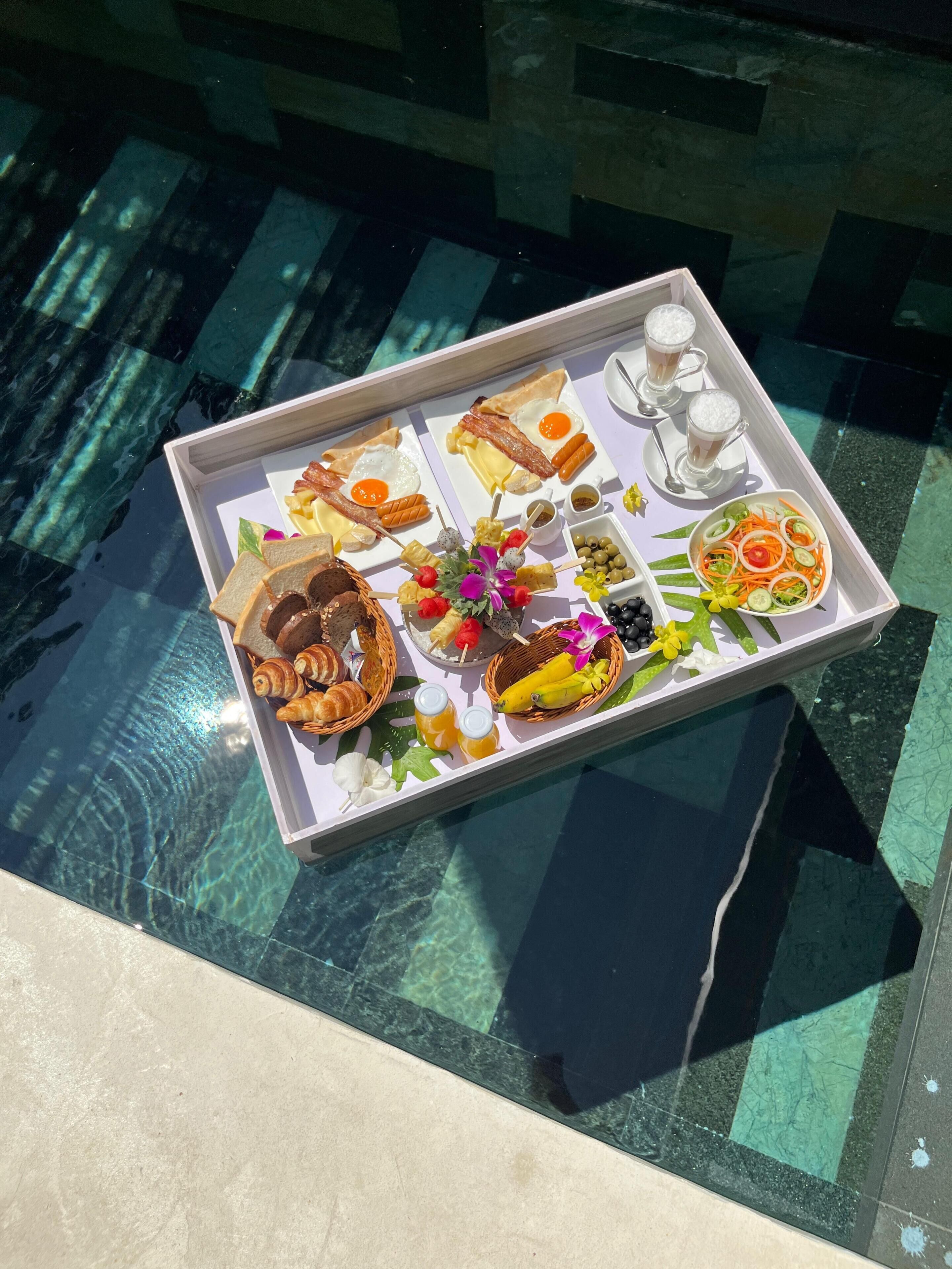 A tray with breakfast items including eggs, bacon, toast, croissants, and fruit, placed on a glass table near a pool.