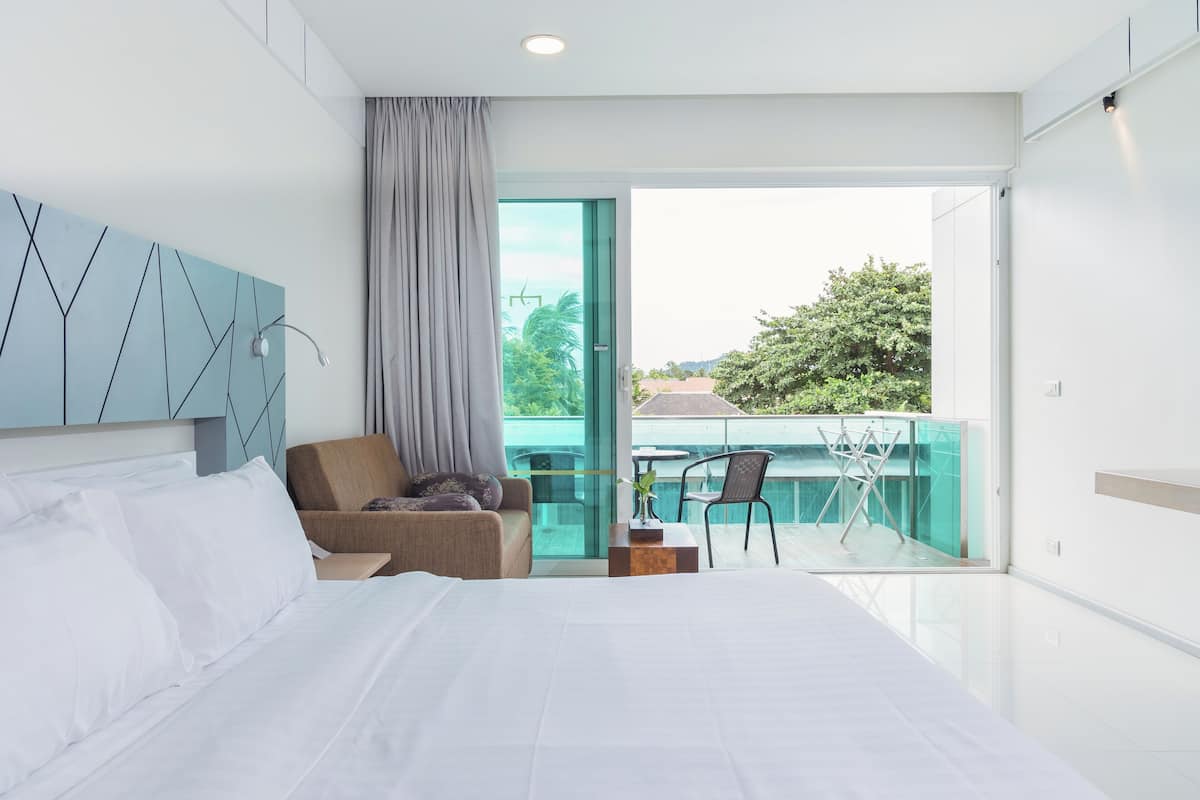 A modern hotel room with a large bed, a sofa, a small table with a plant, and a balcony with a chair and a drying rack.
