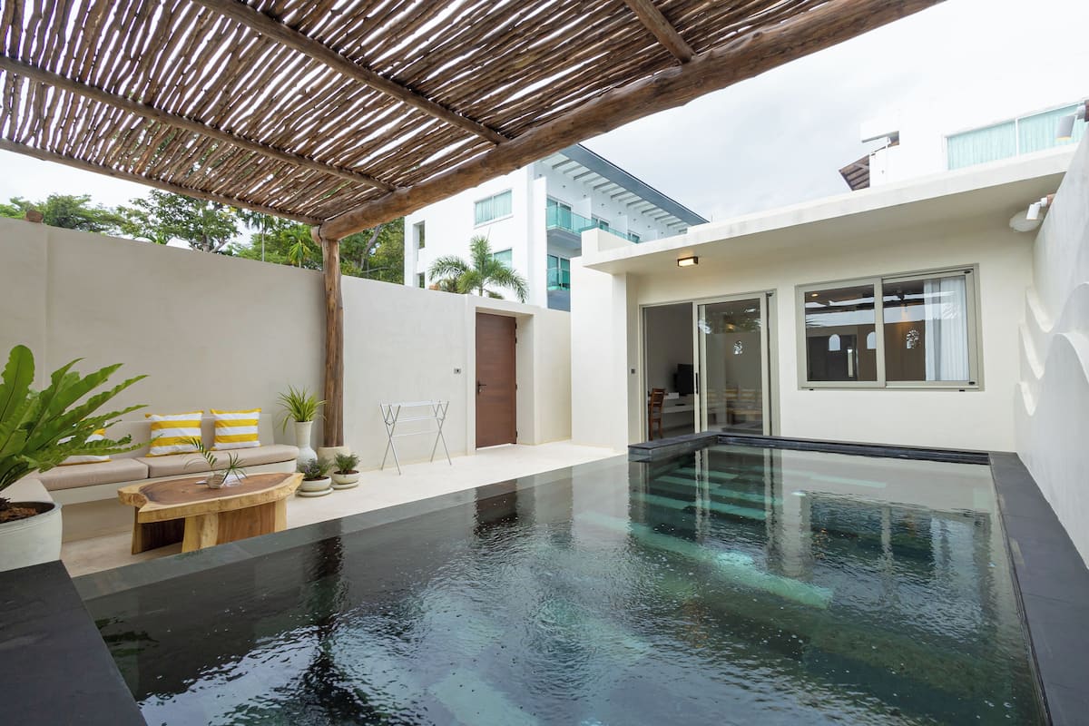 A modern outdoor pool area with a wooden deck, a seating area with cushions, and a building with large windows.