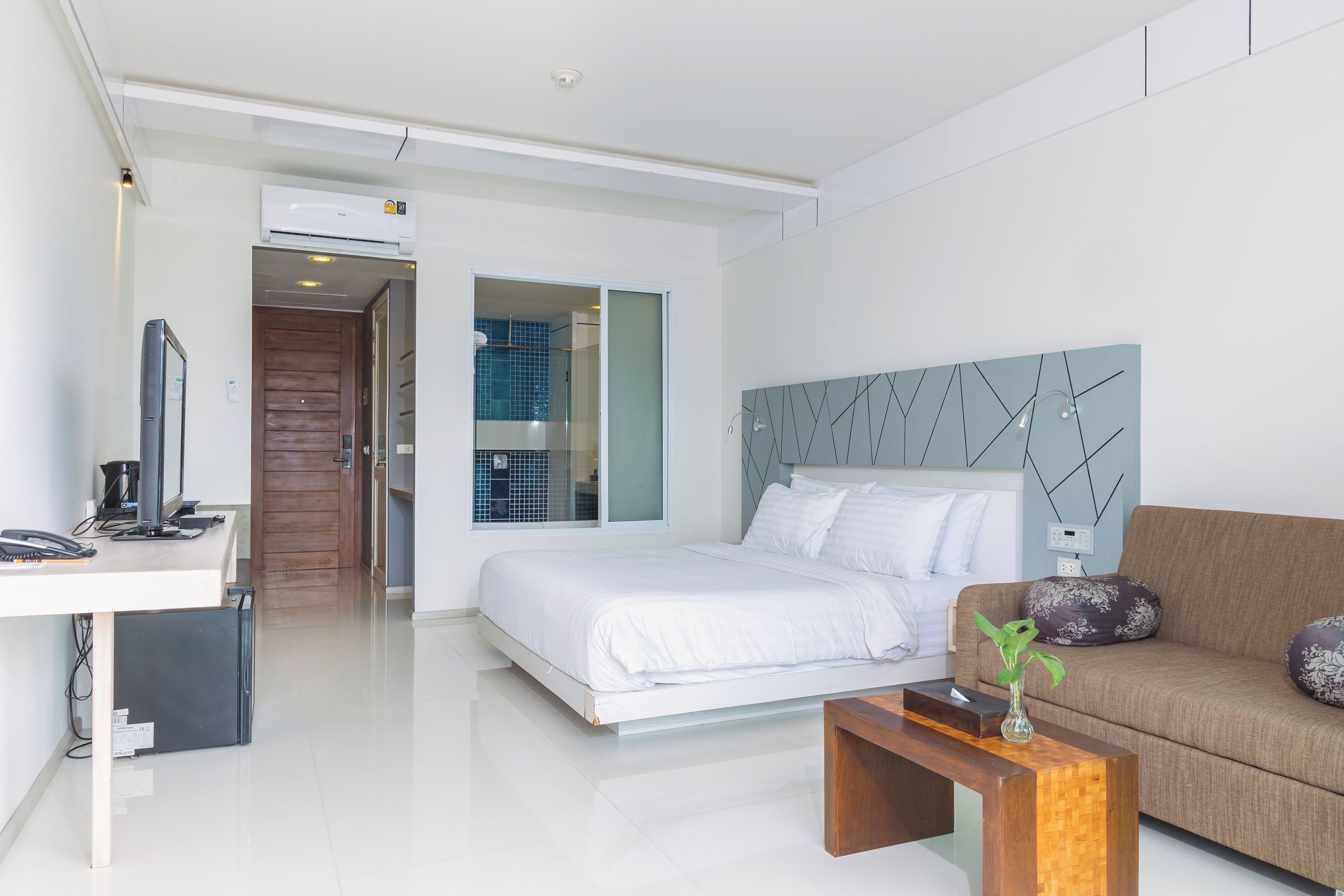 A modern hotel room with a bed, a desk with a computer, a sofa, and a decorative plant.