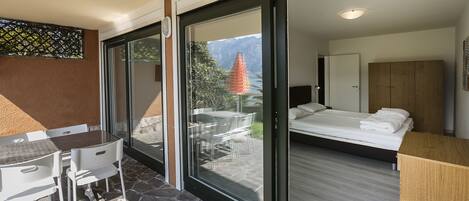Una habitación moderna con una cama, un armario de madera, un área de comedor con una mesa y sillas, y un gran ventanal corredero que da a un patio exterior.