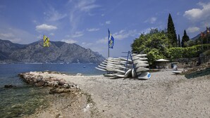 Una playa rocosa con barcos y banderas, agua cristalina y montañas al fondo.