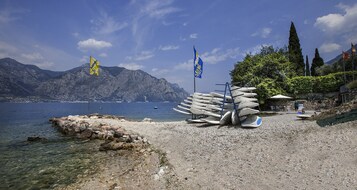 Una playa rocosa con barcos y banderas, agua cristalina y montañas al fondo.