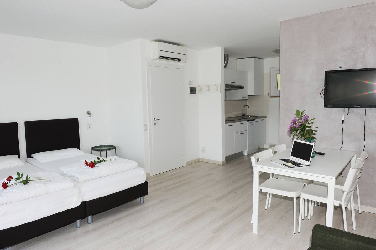 A modern hotel room with a bed, a desk with a laptop, a TV, and a small kitchen area.