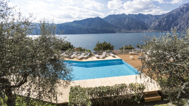 Una piscina con sillones de descanso, rodeada de árboles, con vistas a un lago y montañas.