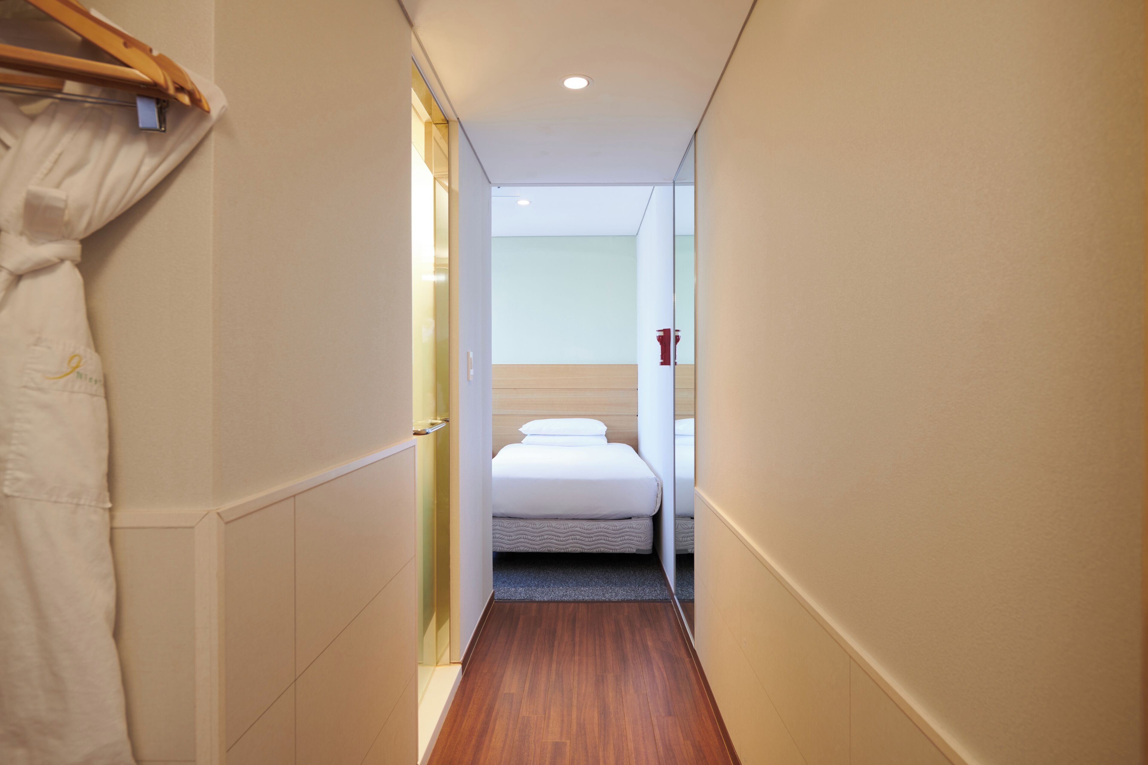 A narrow hallway leading to a bedroom with a bed, a wooden floor, and a white curtain.