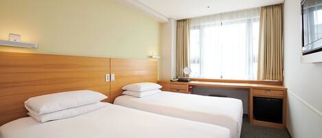 A hotel room with two beds, a desk, a small refrigerator, and a window with curtains.